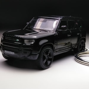 May include: A black die-cast model of a Land Rover Defender SUV, presented as a keychain. The miniature vehicle features detailed design elements, including the Defender logo and a license plate. The wheels and body are all black.