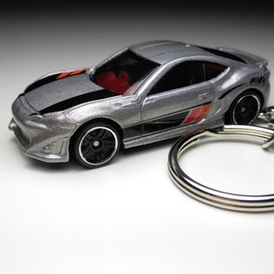 May include: A silver toy car with black and red racing stripes, attached to a silver key ring. The car has black wheels and a black interior.