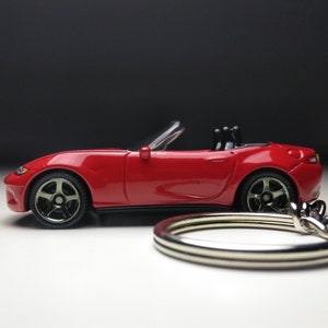 Puede incluir: Un coche de juguete rojo convertible con interior negro y un llavero plateado. El coche tiene una parrilla negra y ruedas negras.