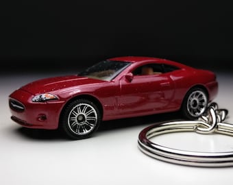 May include: A red toy car keychain featuring a detailed model of a Jaguar XK. The car has silver wheels and a silver keychain ring.