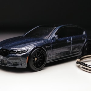 Op de afbeelding: Een miniatuur donkerblauwe auto met zwarte wielen en getinte ramen. De auto heeft een gedetailleerde grille en koplampen. Een zilveren sleutelhanger is aan de auto bevestigd. De auto is een verzamelobject.