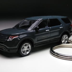 Puede incluir: Un modelo de SUV en miniatura de color gris oscuro con ruedas plateadas y un llavero. El coche de juguete tiene detalles, incluyendo faros y una parrilla delantera. El llavero es de metal plateado.