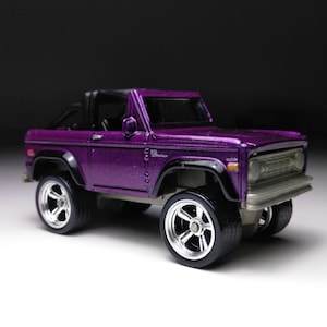 Puede incluir: Modelo de coche de juguete de fundición a presión de color púrpura, que representa un Ford Bronco. El coche tiene detalles en negro y ruedas plateadas.