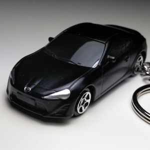 Może przedstawiać: Czarny brelok do kluczy z modelem samochodu odlewanego ciśnieniowo. Miniaturowy samochód ma elegancki design ze srebrnymi kołami i reflektorami. Brelok zawiera srebrny metalowy pierścień.