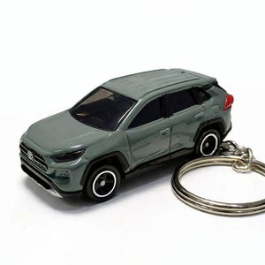 May include: A small, olive-green SUV model with black wheels and a silver keyring. The toy car features detailed design elements such as headlights, taillights, and a front grille. The keyring is made of silver-coloured metal.
