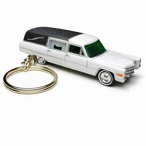 May include: A white miniature hearse keychain with a black roof and green tinted windows. The toy car has silver wheels and a gold-colored keyring. The hearse is a classic car design.