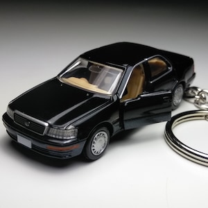 以下が含まれることがあります： 銀色のキーリングが付いた黒いおもちゃの車のキーホルダー。車はドアが開いており、タン色の内装です。