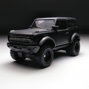 Pode incluir: Um carro de brinquedo Ford Bronco preto com rodas pretas e a palavra "Bronco" na grelha dianteira.