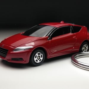 Puede incluir: Un coche rojo en miniatura con detalles en negro y plata. El coche de juguete está unido a un llavero plateado. El coche tiene un diseño elegante con faros y ruedas visibles. El fondo es un degradado de blanco y negro.