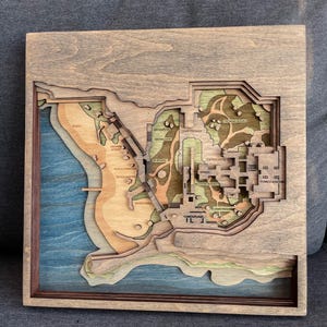 May include: A multi-layered wooden map with a detailed topographical design. The map features a coastal area with blue water, tan beaches, and green and brown landmasses. Text labels identify locations. The frame is a dark brown color.