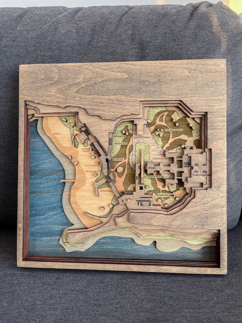 May include: A multi-layered wooden map with a detailed topographical design. The map features a coastal area with blue water, tan beaches, and green and brown landmasses. Text labels identify locations. The frame is a dark brown color.