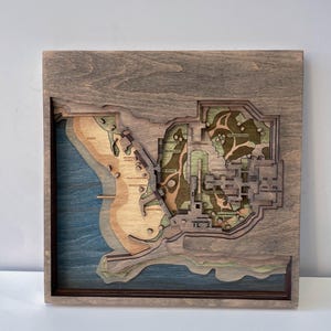 May include: A wooden, layered map featuring a coastal landscape with beach, water, and architectural details. The map is framed in a dark wood, with the map itself showcasing various shades of brown, green, and blue. The map includes text labels.