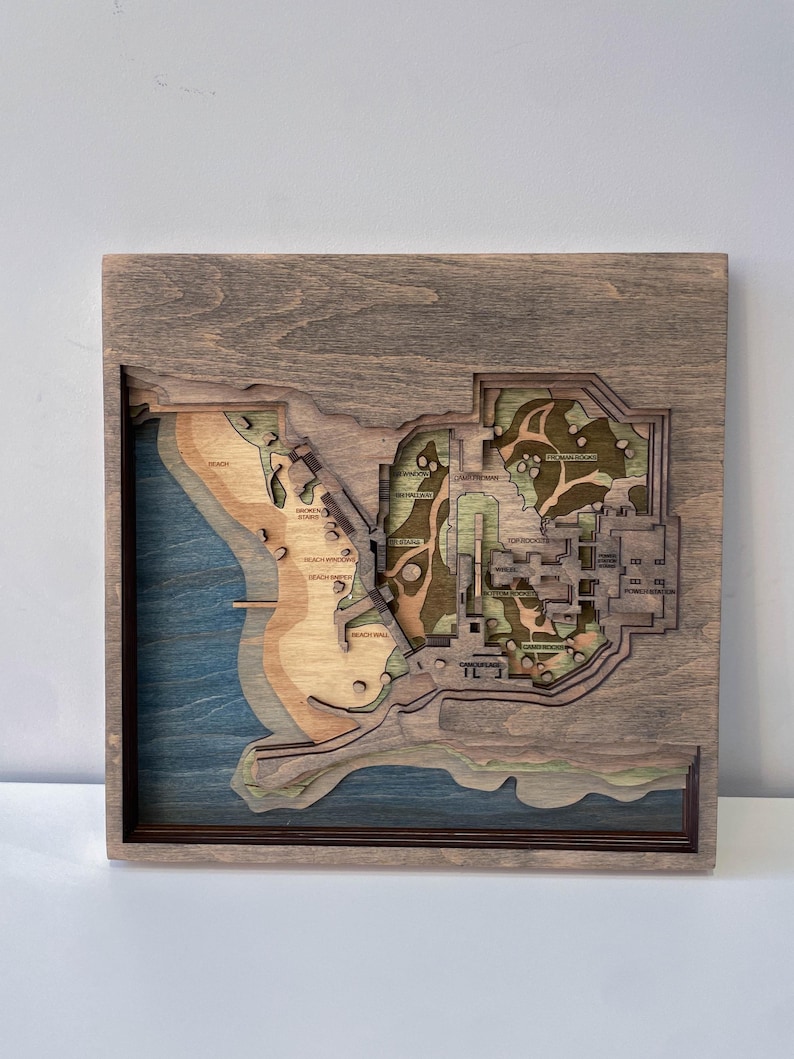 May include: A wooden, layered map featuring a coastal landscape with beach, water, and architectural details. The map is framed in a dark wood, with the map itself showcasing various shades of brown, green, and blue. The map includes text labels.