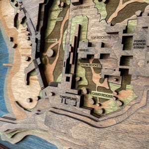 May include: A layered wooden map with a detailed topographical design. The map features various terrains, including beaches, hills, and structures, with text labels such as "Camp Froman" and "Top Rockets." The color palette includes shades of brown, green, and blue.