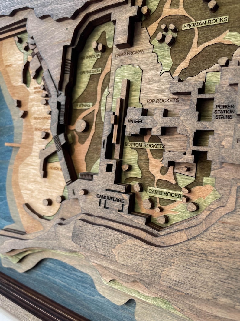 May include: A layered wooden map with a detailed topographical design. The map features various terrains, including beaches, hills, and structures, with text labels such as "Camp Froman" and "Top Rockets." The color palette includes shades of brown, green, and blue.