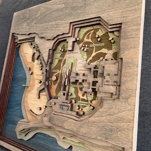 May include: A layered wooden map with a gray frame. The map features a detailed topographical design with various levels and colors, including blue, green, and brown. Text labels like "Power Station" and "Camo Rocks" are visible.