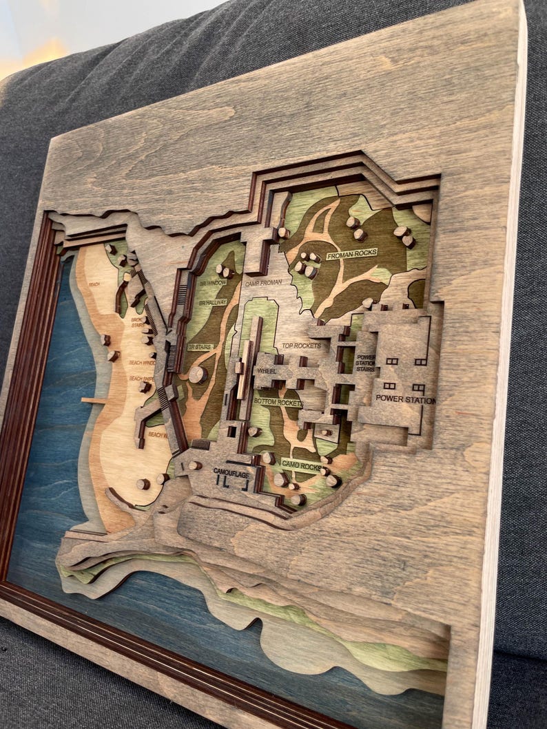 May include: A layered wooden map with a gray frame. The map features a detailed topographical design with various levels and colors, including blue, green, and brown. Text labels like "Power Station" and "Camo Rocks" are visible.