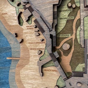 May include: A multi-layered wooden map with a beach and building design. The map features text labels such as "BEACH", "BROKEN STAIRS", "BEACH WINDOWS", "BEACH SNIPER", "BEACH WALL", "BR WINDOW", "BR HALLWAY", and "BR STAIRS". The map is made of various shades of brown, green, and blue.