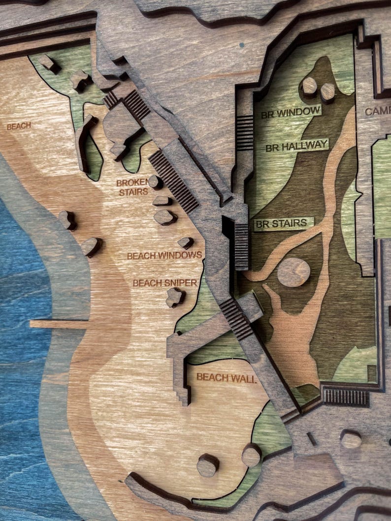 May include: A multi-layered wooden map with a beach and building design. The map features text labels such as "BEACH", "BROKEN STAIRS", "BEACH WINDOWS", "BEACH SNIPER", "BEACH WALL", "BR WINDOW", "BR HALLWAY", and "BR STAIRS". The map is made of various shades of brown, green, and blue.