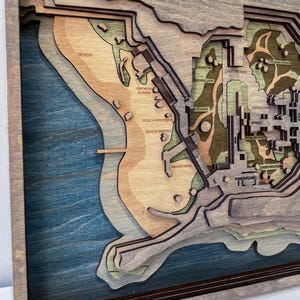 May include: A layered wooden map with a coastal theme, featuring a beach, stairs, and buildings. The map uses shades of blue, brown, and green, with text labels like "Beach" and "Broken Stairs". The map is framed in a dark brown border.