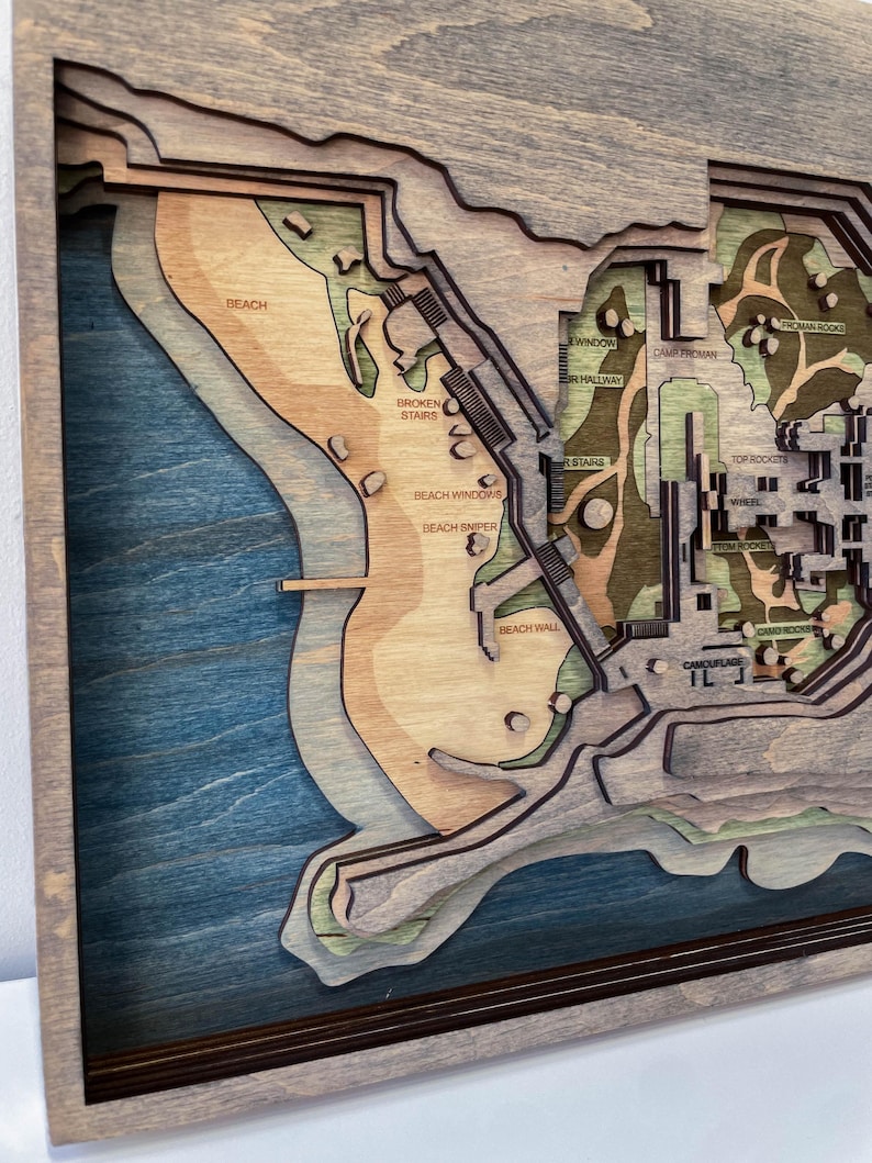 May include: A layered wooden map with a coastal theme, featuring a beach, stairs, and buildings. The map uses shades of blue, brown, and green, with text labels like "Beach" and "Broken Stairs". The map is framed in a dark brown border.