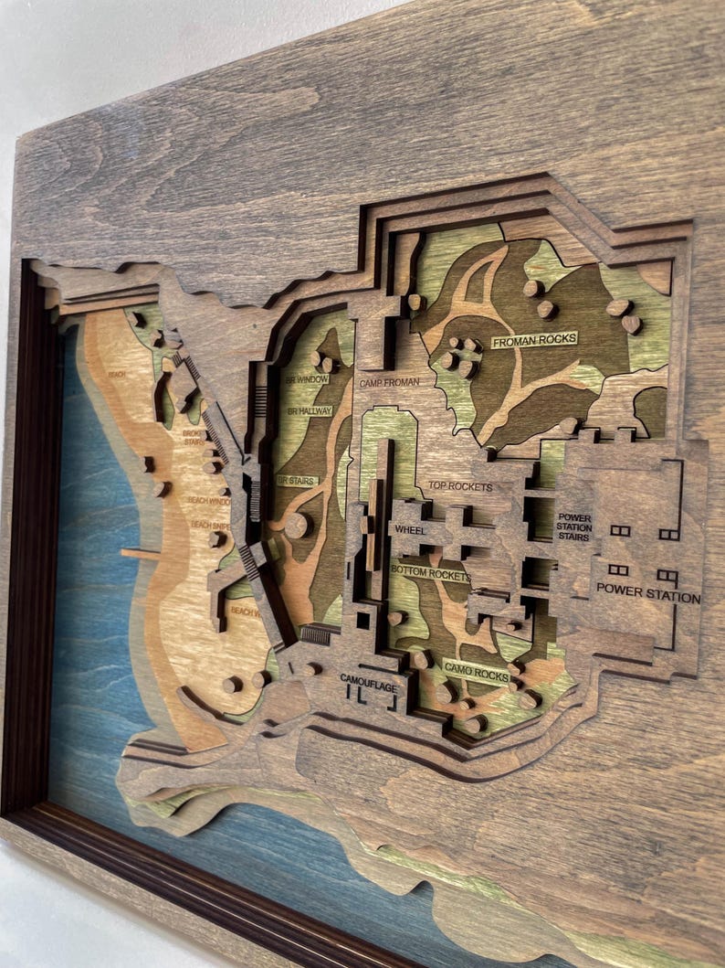 May include: A layered wooden map with a dark brown frame. The map features a detailed topographical design with various colors, including shades of brown, green, and blue. Text labels such as "FROMAN ROCKS" and "POWER STATION" are visible.