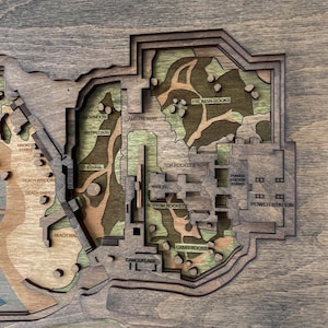 May include: A multi-layered wooden map with a detailed layout. The map features text labels such as "FROMAN ROCKS", "POWER STATION", and "CAMOUFLAGE". The color palette includes shades of brown, green, and blue, creating a topographic effect.