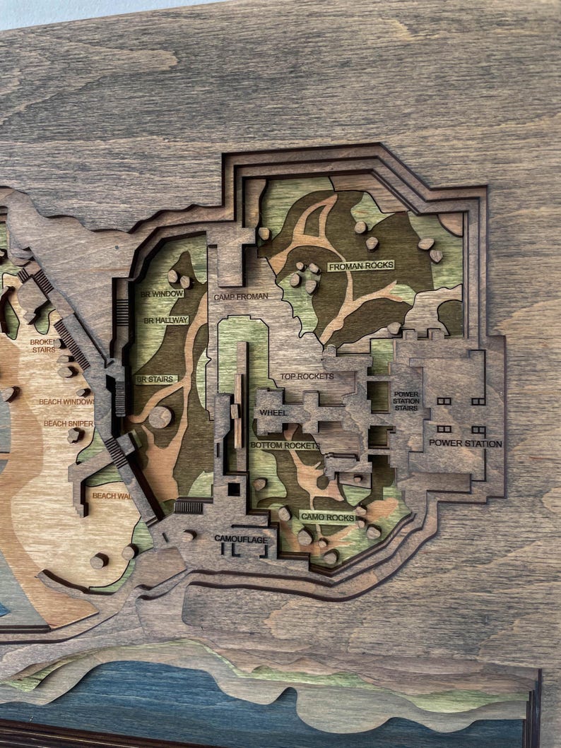 May include: A multi-layered wooden map with a detailed layout. The map features text labels such as "FROMAN ROCKS", "POWER STATION", and "CAMOUFLAGE". The color palette includes shades of brown, green, and blue, creating a topographic effect.