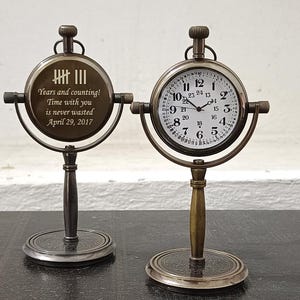 May include: Two antique-style brass desk clocks on stands. One clock face shows numbers 1-24 in black on a white background. The other side features an engraved tally mark design and the text: Years and counting! Time with you is never wasted April 29, 2017.