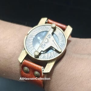 May include: A brass sundial watch featuring a circular face with engraved Roman numerals and a central gnomon displaying a sun and moon icon. The timepiece is attached to a tan leather strap secured with two brass rivets. The text AliHasnainCollection is visible.