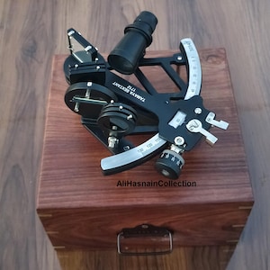 May include: A black Tamaya Sextant 1712 navigational instrument with a silver arc and adjustment knobs, resting on a wooden box. The box has a metal handle and is made of dark wood. The text "AliHasnainCollection" is visible.
