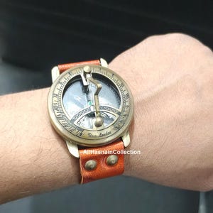 May include: A brass sundial watch with a brown leather strap featuring two metal studs. The circular face displays Roman numerals, a central gnomon, and the text Ross London. The compass dial is visible beneath the gnomon, showing cardinal directions.
