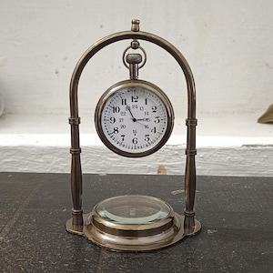 May include: A brass-toned metal desk clock suspended within an arched frame. The clock face is white with black numerals from 1 to 24, featuring a smaller inner ring of numbers and a circular glass base component at the bottom of the stand.