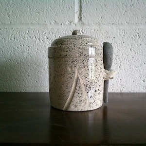 May include: A white ceramic jar with black speckles and a wooden spoon attached to the side. The jar has a lid and is sitting on a dark wooden surface.