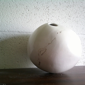 May include: A white ceramic vase with a round, spherical shape. The vase has a small opening at the top and a subtle brown line running down the side.