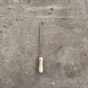 Peut inclure: Un outil de crochetage de tapis en métal doté d'une longue tige en acier fine avec un crochet courbé à l'extrémité supérieure et un manche en bois clair lisse à la base, relié par une petite virole en métal argenté pour une prise en main.