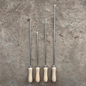 Peut inclure: Un ensemble de quatre outils en métal avec des manches en bois clair. Les outils présentent de longues tiges fines de longueurs variables. Un outil possède un crochet courbé à l'extrémité, tandis que les trois autres ont des pointes plates.