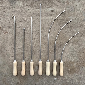 Peut inclure: Un ensemble de sept outils en métal avec des manches en bois clair. La collection comprend un outil à crochet, deux outils à pointe droite et quatre outils à pointe courbée de longueurs variées, tous dotés de poignées en bois cylindriques.