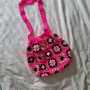 May include: A pink and black crocheted tote bag with a long pink strap. The bag is made of granny squares.