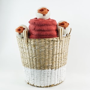 Peut inclure: Un panier en osier avec une base blanche contient une serviette rouge pliée et trois boules de séchage en laine blanche avec des motifs de moutons bruns. Deux pinces à linge en bois fixent les boules de séchage et la serviette. Le panier est en matériau tissé naturel et blanc.