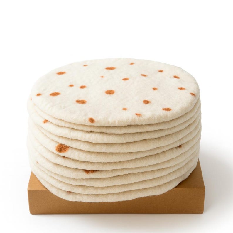 Puede incluir: Una pila de tortillas blanquecinas con manchas marrones, sobre una peque&ntilde;a caja de cart&oacute;n marr&oacute;n. Las tortillas son redondas y parecen suaves, con una superficie ligeramente texturizada. La imagen est&aacute; sobre un fondo blanco.