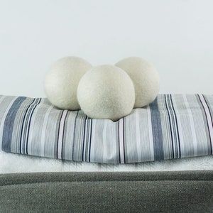 May include: Three white wool dryer balls rest on a folded striped fabric, atop a stack of towels inside a gray storage basket. The fabric has blue, gray, and burgundy stripes. The basket has a gray handle.