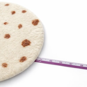 Puede incluir: Un salvamanteles redondo de fieltro blanquecino con manchas marrones, que se asemeja a una tortilla. Una cinta m&eacute;trica morada se extiende por la imagen, mostrando medidas en cent&iacute;metros. El salvamanteles tiene aproximadamente 35,5 cm de di&aacute;metro.