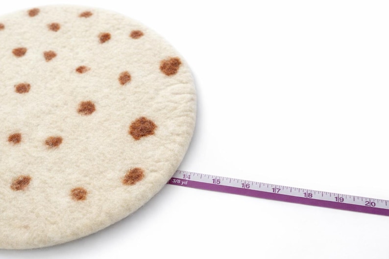 Puede incluir: Un salvamanteles redondo de fieltro blanquecino con manchas marrones, que se asemeja a una tortilla. Una cinta m&eacute;trica morada se extiende por la imagen, mostrando medidas en cent&iacute;metros. El salvamanteles tiene aproximadamente 35,5 cm de di&aacute;metro.