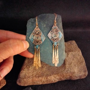 Peut inclure: Une paire de boucles d'oreilles en cuivre avec une patine turquoise. Chaque boucle d'oreille présente un pendentif en forme de diamant avec un motif en spirale et quatre brins pendants.