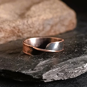 Può includere: Un anello in rame e argento con un design attorcigliato. L'anello è su una superficie di pietra grigio scuro.