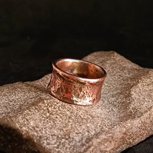 May include: A wide copper band ring with a hammered texture. The ring is sitting on a brown rock.