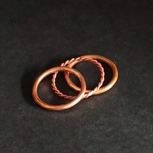 May include: Three copper rings, one with a twisted design, stacked on top of each other against a black background.
