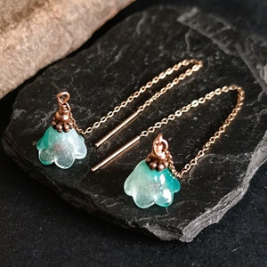 Puede incluir: Un par de aretes delicados con un diseño de flor turquesa y una cadena dorada. Los aretes están sobre una superficie de pizarra negra.