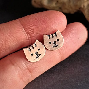 Peut inclure: Une paire de boucles d'oreilles clous en cuivre en forme de visage de chat. Les boucles d'oreilles sont petites et ont un design simple.